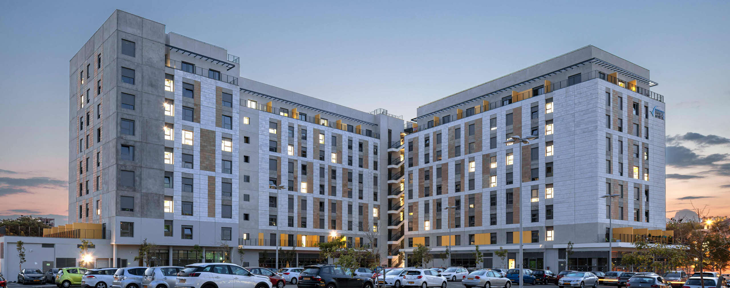 בניין מגורים מודרני עם שמונה קומות בתאורה לילית ושורת מכוניות חונות בחזית.