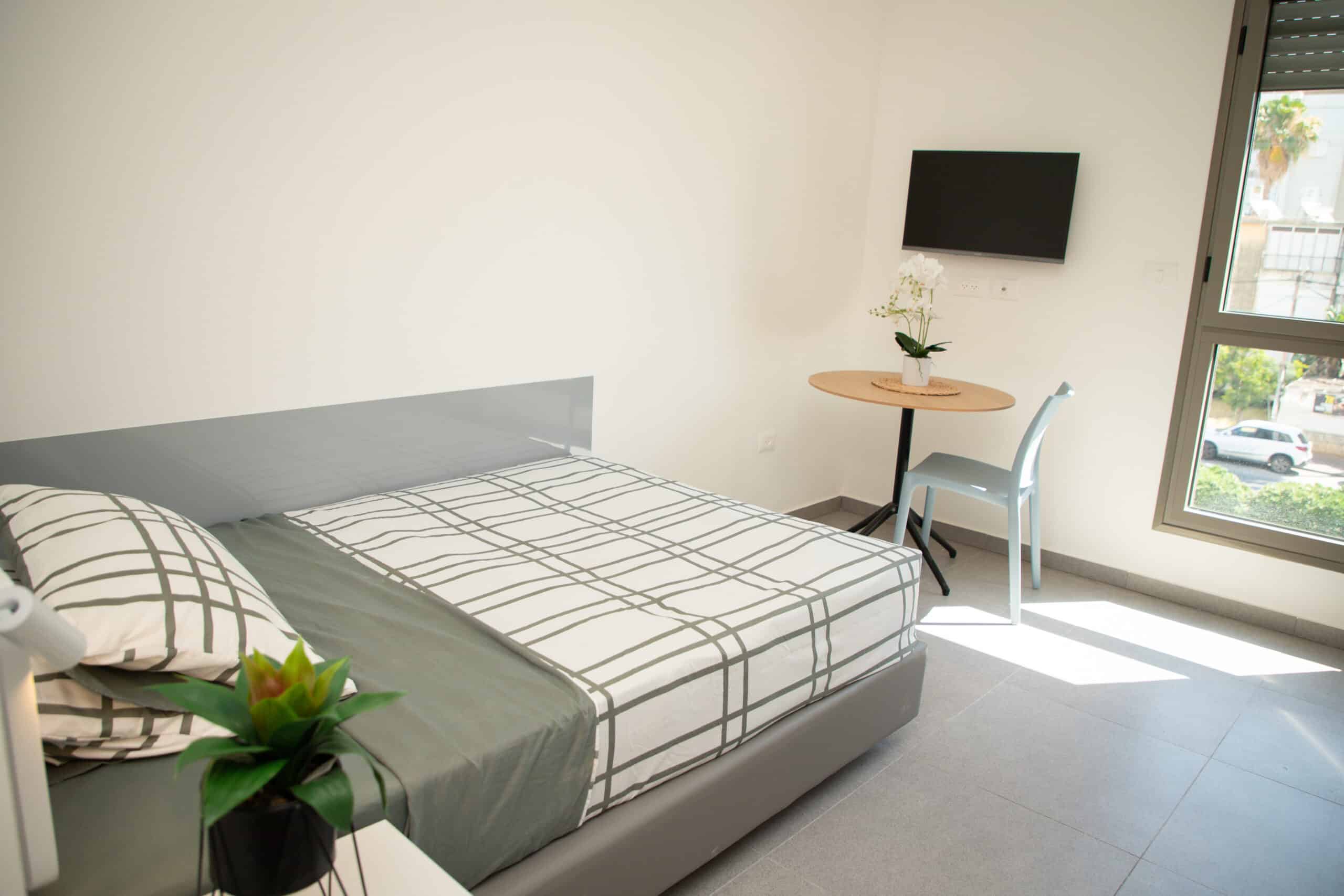 חדר שינה מודרני עם מיטה בעלת מצעים בדוגמת ריבועים, טלוויזיה תלויה על הקיר, שולחן קטן עם כיסא, וצמח דקורטיבי.