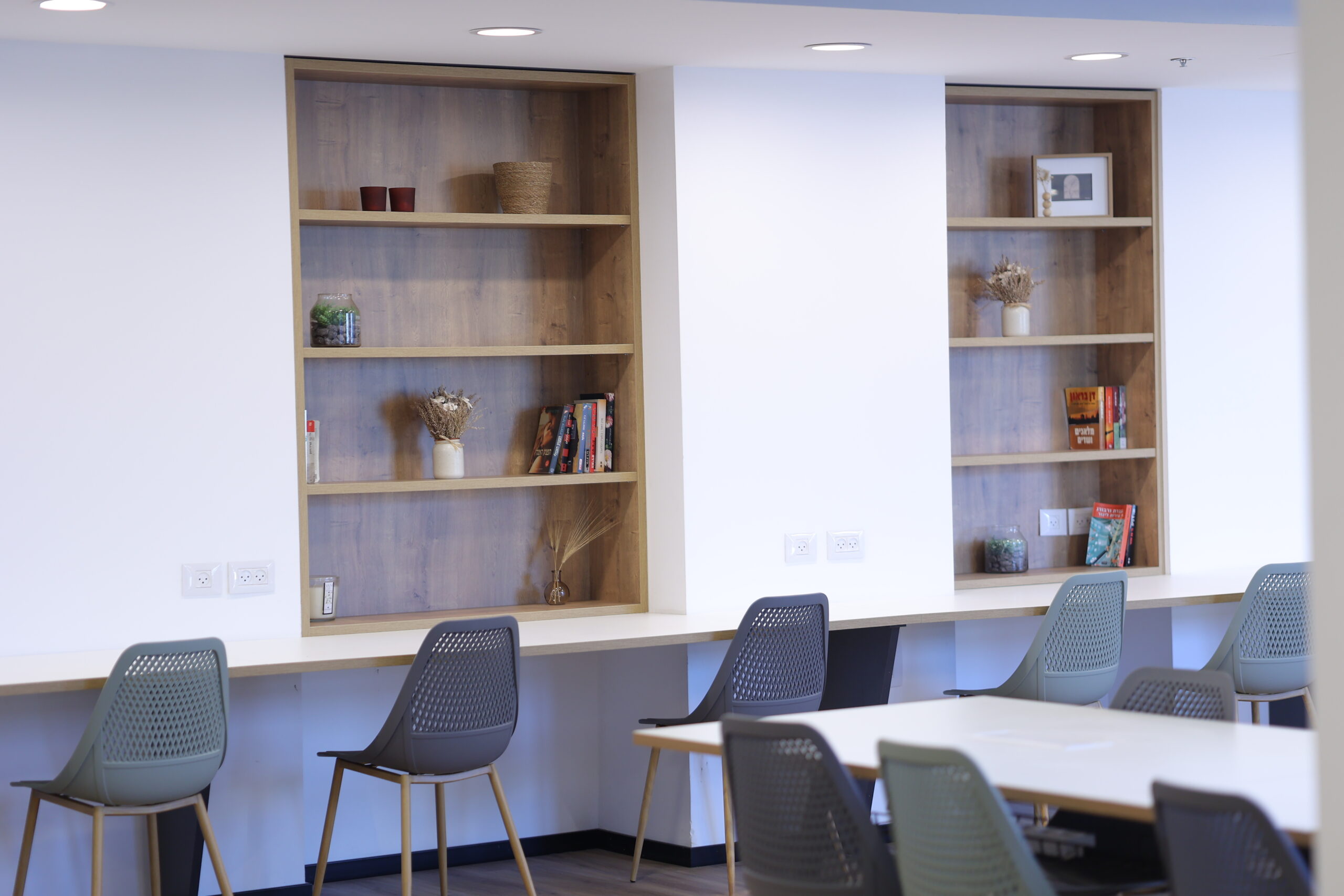 פינת עבודה עם כיסאות פלסטיק אפורים ושולחן עבודה ארוך עם מדפים מעוצבים על הקיר המכילים ספרים, צמחים וקישוטים.