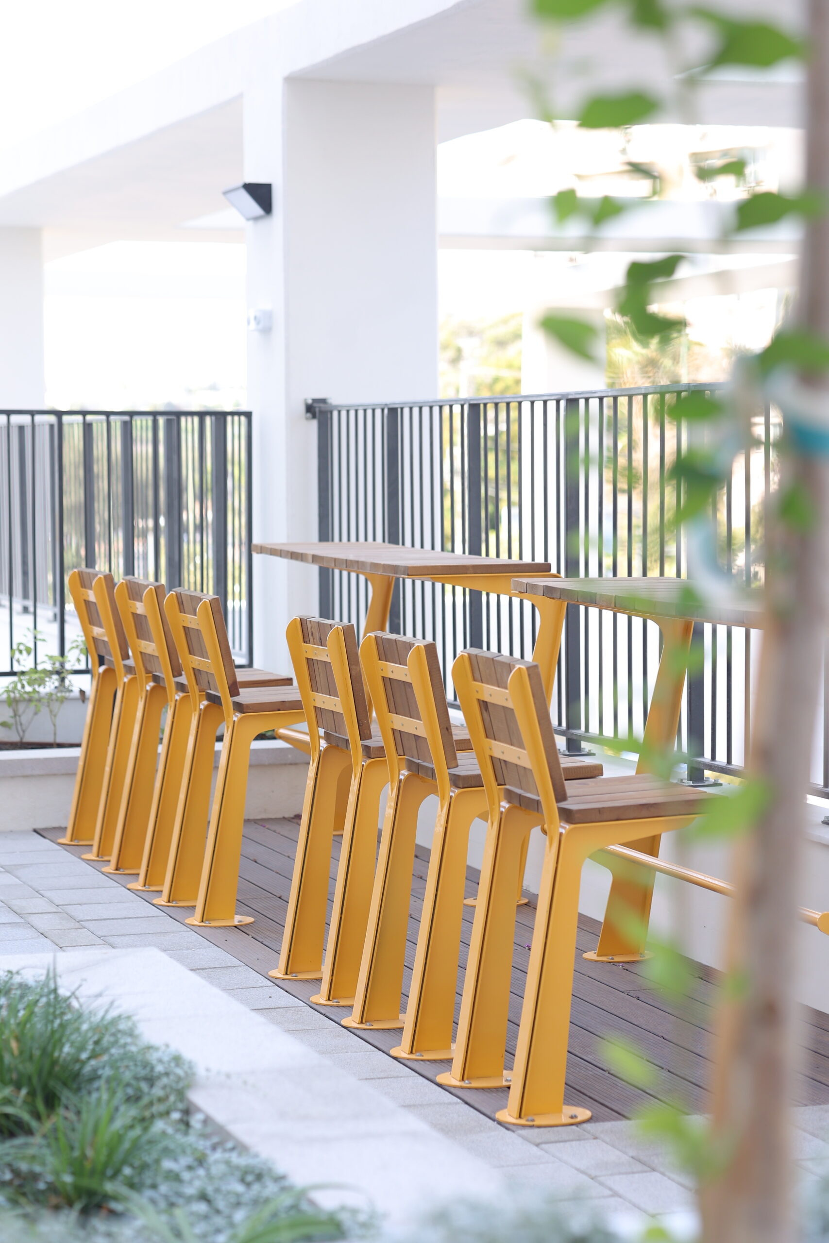 שולחן גבוה עם כיסאות עץ מתכת בצבע צהוב, מסודרים בשורה ארוכה על מרפסת עם מעקה מתכת, צמחייה משתקפת מלפנים.
