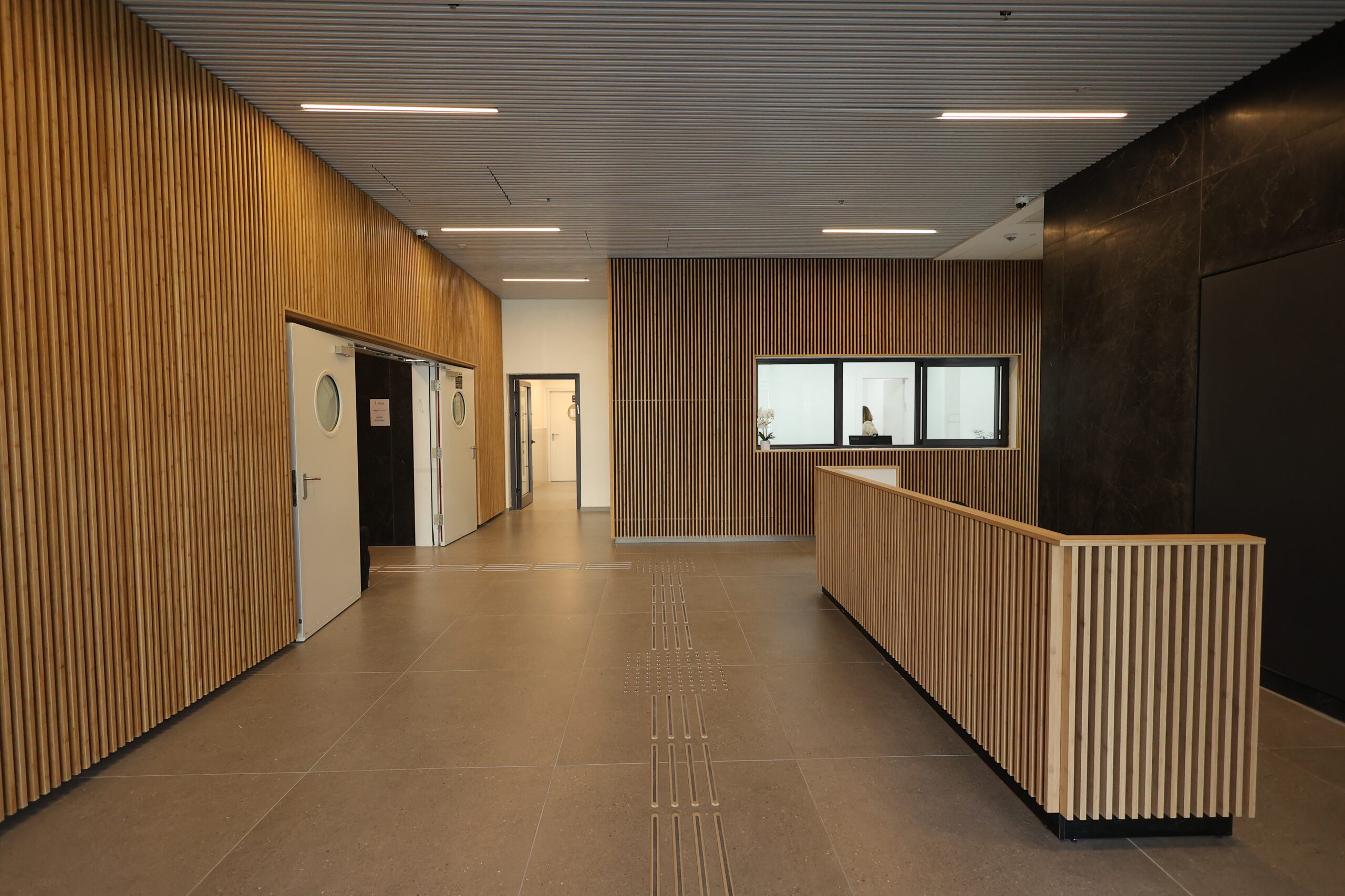 לובי מודרני עם קירות ותקרה בציפוי עץ, דלפק קבלה מעץ, דלתות לבנות עם חלונות עגולים ואדם יושב מאחורי דלפק זכוכית.